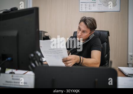 Office worker reading a document while talking on a landline phone. Multitasking, heavy workload, and focused business routine. Stock Photo