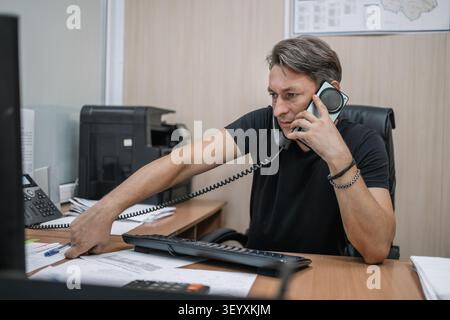 Businessman multitasking on a phone call while reaching for papers and a pen at his office desk. High workload and busy day. the man is talking on two Stock Photo