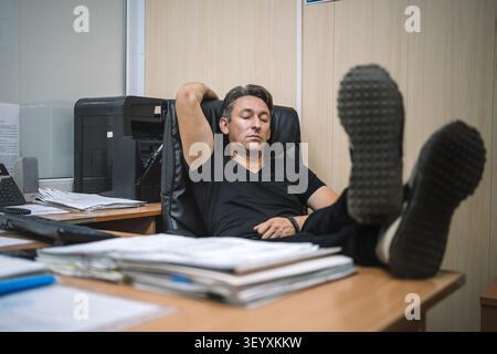 Tired office worker resting with his feet up on the desk, eyes closed. Work burnout, relaxation break, and office fatigue. Stock Photo