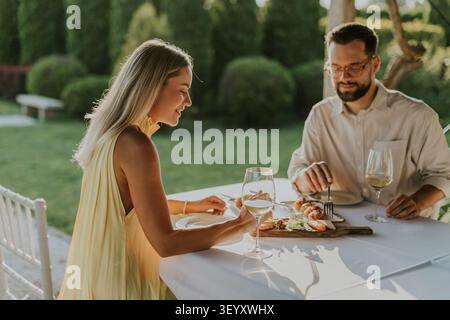 A woman and man sit at a table in a garden during evening. They are having dinner, with a platter of food in front of them. They drink wine and engage Stock Photo