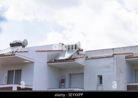 Two solar thermal water heaters installed on the flat roof of a white building under a bright cloudy sky. The structure features terracotta tiles and Stock Photo