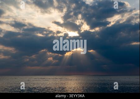 The sun shines out from behind thick clouds over the sea in the late afternoon. Stock Photo