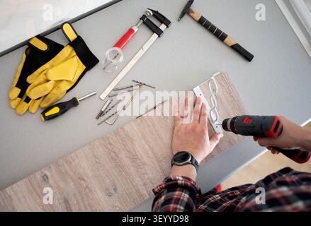 Person drills white metal bracket onto wood plank with power drill. Tools lay on gray table. Stock Photo