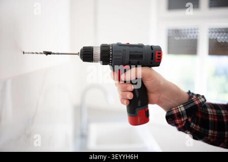 Adult man drills hole in white tiled kitchen wall. Wears plaid shirt, black cap. Stock Photo