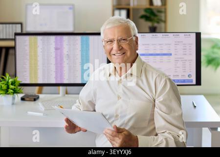Smiling Senior Accountant Reviewing Reports on Computer with Spreadsheets in Office Stock Photo