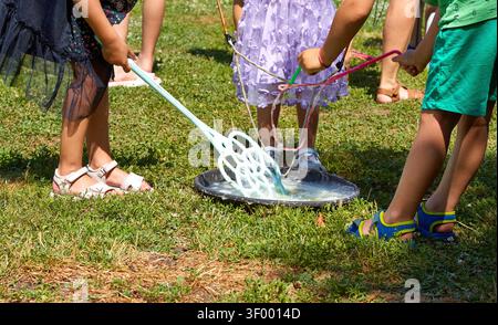 Children gather on green grass, using colorful wands to catch soap bubbles from a large bowl of bubble solution in a sunny outdoor park Stock Photo