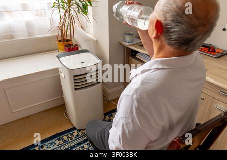 An elderly man cooling down in front of a portable air conditioner and drinking water during a Summer heat wave. Stock Photo