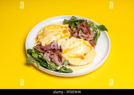 Close-up of Eggs Benedict topped with creamy hollandaise sauce, served with ham and greens on a bright yellow background. Perfect for restaurant menus Stock Photo