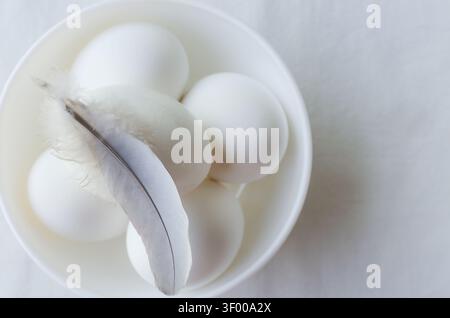 Five white chicken eggs lie in a white bowl and a light feather lies on top of them on a white background. Egg concept. From farm to table. Minimalist Stock Photo