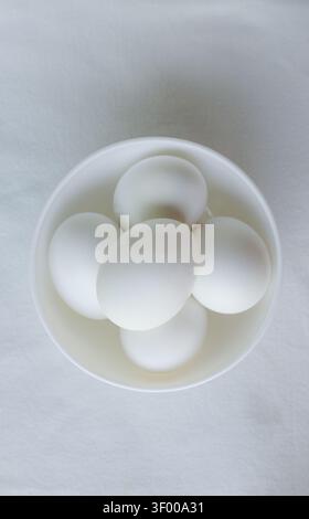 Five white chicken eggs lie in a white bowl on a white background. Egg concept. From farm to table. Clean monochrome style, emphasis on simplicity, te Stock Photo