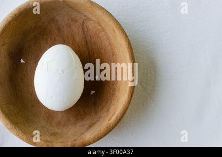 White egg with cracked shell in the center of a wooden bowl. Minimalistic composition - the smooth fragility of the egg and the rough texture of the w Stock Photo