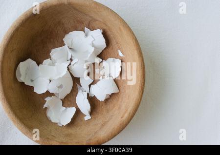 Wooden bowl with fragments of white egg shells on a light background. Minimalist still life composition, contrast between smooth white forms and warm Stock Photo