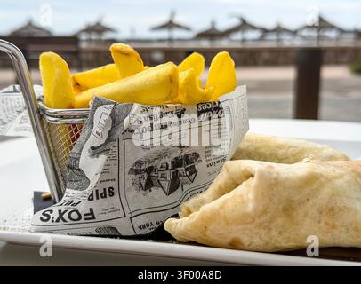 A plate of wraps & chips served in a metal basket with newspaper wrapping at an outdoor seaside cafe Stock Photo