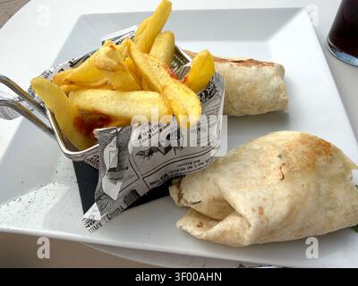 A white plate served with two tortilla wraps & golden French fries in a mini wire basket lined with faux newspaper print Stock Photo