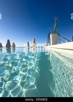 Water level view of an infinity swimming pool with closed umbrellas and blue sky Stock Photo