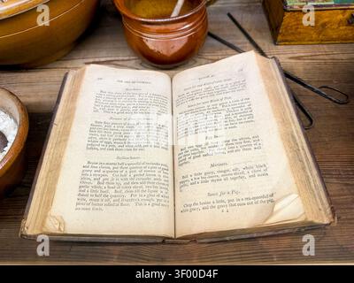 Open pages of The Art of Cookery, an old cookbook displayed on a wooden table with rustic pottery & kitchen utensils Stock Photo