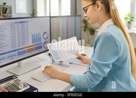 Businesswoman analyzing finance dashboard and reports on computer screens at desk Stock Photo