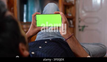 An over-the-shoulder, eye-level shot of a young man relaxing at home while lying down and holding a mobile smartphone horizontally with a green screen. Stock Photo