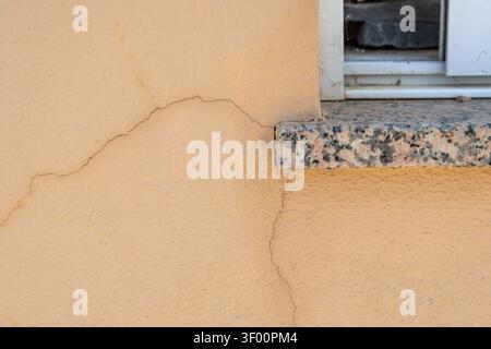 Close-up view of hairline cracks on a textured beige exterior wall adjacent to a speckled granite window ledge and white window frame Stock Photo