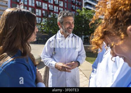 Healthcare professionals, including a male doctor and two female colleagues, engaging in a smiling conversation outdoors during a sunny day, represent Stock Photo