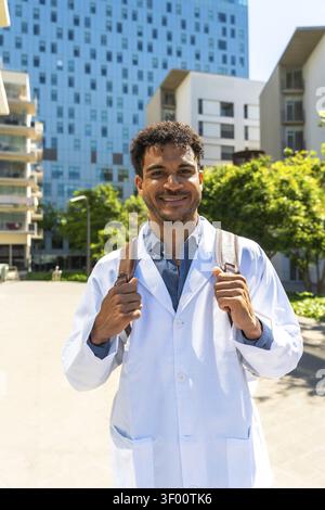 Young black male medical student or doctor smiling and looking directly at the camera, wearing a lab coat and backpack on a university campus or hospi Stock Photo