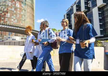 Diverse team of doctors and nurses walking outdoors with coffee and charts, smiling and talking near a modern hospital healthcare teamwork, collaborat Stock Photo