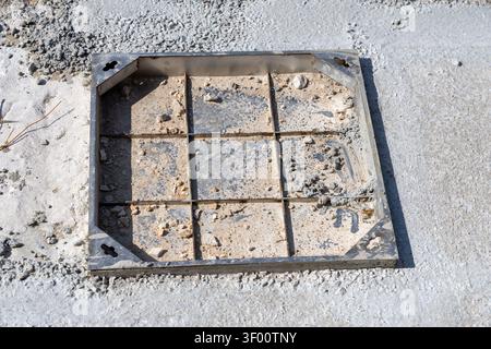A close-up view of a square metal grate cover, partially filled with dirt and debris, set into a rough concrete surface at a building site Stock Photo