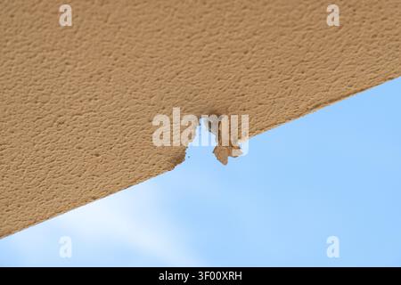 A section of beige stucco is peeling away, revealing a hole and the bright blue sky beyond, suggesting damage or wear Stock Photo