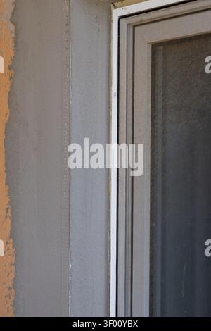 Detailed texture of a rough plaster wall adjacent to a white window frame and mesh screen, showing subtle imperfections and construction elements Stock Photo