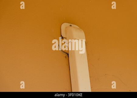 A close-up view shows a cracked and deteriorating white plastic downspout secured to a rough, ochre-colored exterior wall Stock Photo
