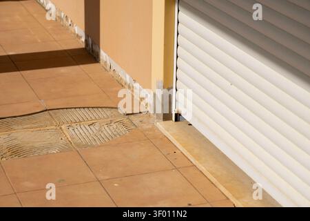 Close-up view of a building's exterior showing a damaged wall base with missing plaster and cracked, dislodged terracotta floor tiles next to a white Stock Photo