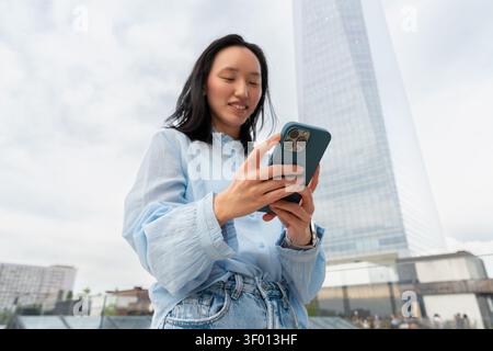 Low angle view of a Gen Z Asian woman smiling while using a smartphone outdoors, casual denim outfit, modern city skyline and tall skyscraper in the b Stock Photo