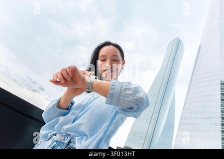 Low-angle view of a Gen Z Asian woman checking her smartwatch outdoors, smiling in a modern business district with glass skyscrapers and bright sky. Stock Photo