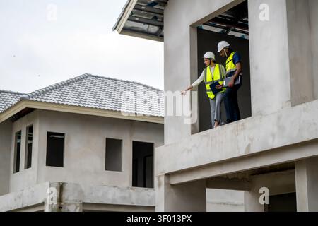 Construction managers holding clipboard and tablet pointing down from second floor balcony of unfinished residential house, concept of teamwork and pr Stock Photo