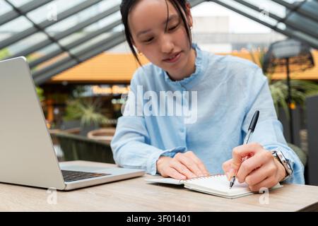 Gen Z asian woman studying at a modern cafe, using a laptop and writing notes in a notebook. Focused student working remotely in a bright indoor space Stock Photo