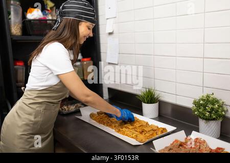 A cafe worker in an apron and striped headscarf is arranging savory pastries on a tray in a catering kitchen. The scene highlights food preparation an Stock Photo