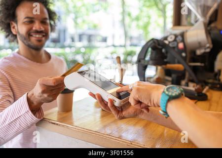 Smiling customer pays by card at a small coffee kiosk counter, holding takeaway drink while contactless terminal processes payment, highlighting small Stock Photo