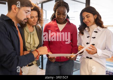 Diverse coworkers gather in a modern coworking office, reviewing printed documents and discussing ideas for a project, showing teamwork, collaboration Stock Photo