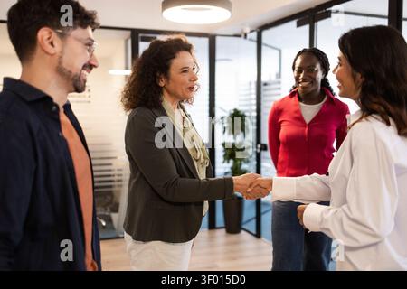 Diverse coworkers meet in a modern coworking office, smiling as two colleagues shake hands to welcome a new team member, reflecting collaboration, tru Stock Photo