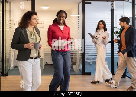 Diverse coworkers walk and talk in a modern coworking office corridor, sharing notes on a project while colleagues chat in the background, reflecting Stock Photo