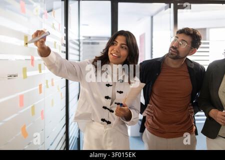 Diverse coworkers brainstorm in a modern office, writing on a glass wall with sticky notes, planning strategy and collaborating on a project during a Stock Photo