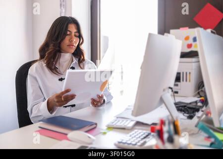 Professional woman sits at a desk in a bright modern office, reading printed documents beside computer monitors and stationery, focused on administrat Stock Photo