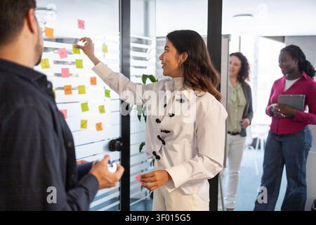 Diverse coworkers brainstorm in a modern office, placing colorful sticky notes on a glass board during a team planning session, sharing ideas, strateg Stock Photo