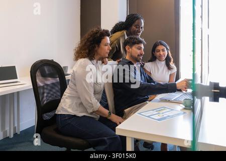 Diverse coworkers gather in a modern coworking office, brainstorming around a laptop and documents. Team members collaborate, discuss ideas, and plan Stock Photo