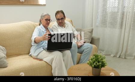 Senior couple sitting on a sofa at home, using a laptop together and discussing online tasks. Relaxed lifestyle scene with technology, learning, and c Stock Photo