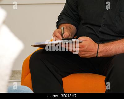 Man writing notes on a clipboard while seated at an indoor event Stock Photo