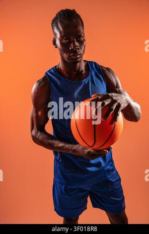 Studio portrait of a black basketball player in blue uniform holding an orange ball, standing against a bright orange background with focused expressi Stock Photo
