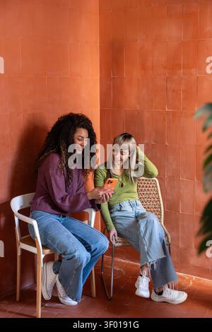Two multiethnic flatmates sit on chairs by a terracotta wall, smiling while looking at a smartphone, sharing a relaxed moment of friendship in their a Stock Photo