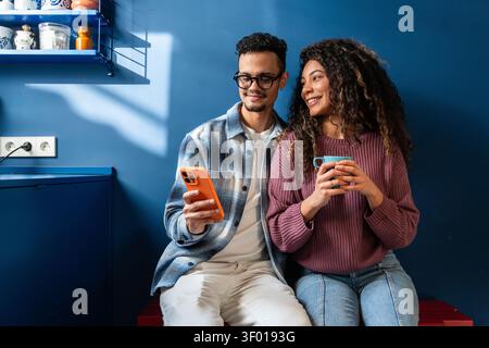 Smiling Latin couple sitting together in a modern blue kitchen, holding a smartphone and a coffee mug, sharing a relaxed moment at home in warm daylig Stock Photo
