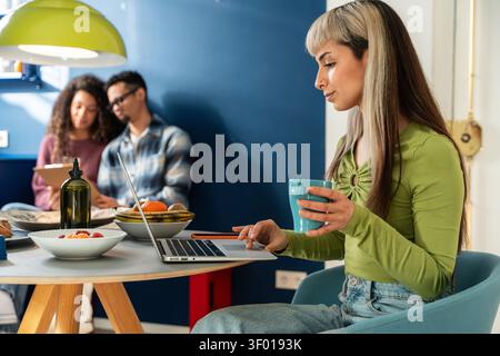 Young woman drinks coffee while working on a laptop at a dining table in a shared apartment, with multiethnic flatmates chatting in the background. Stock Photo
